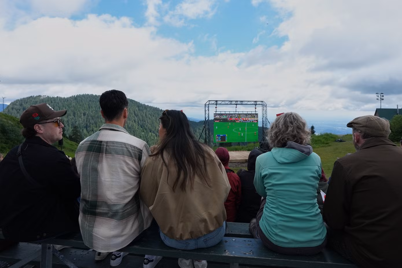 Phía sau màn hình đang chiếu trận đấu World Cup là không gian bao la, với mây trời và các đỉnh núi.