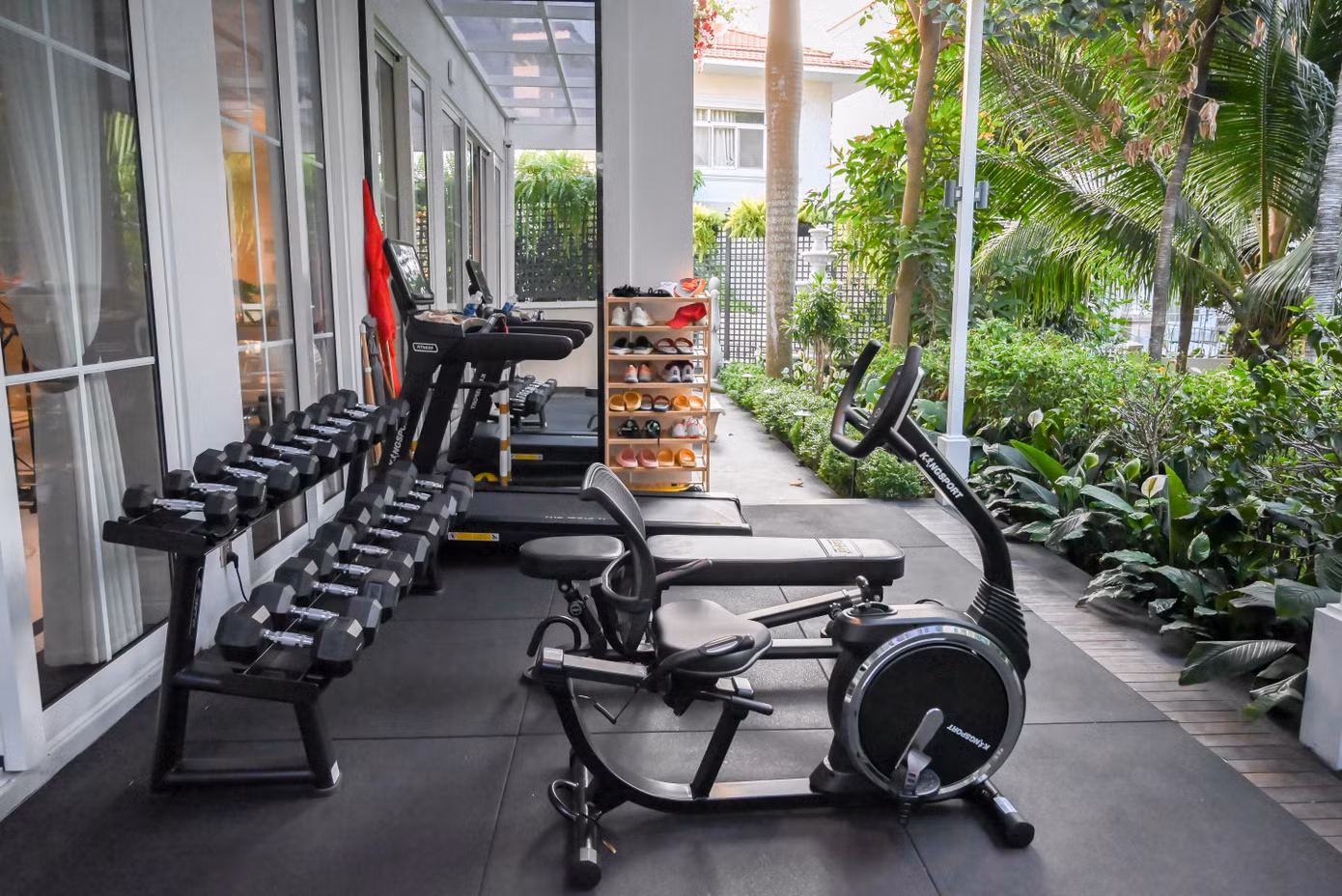 Thay vì bố trí phòng gym kín, gia đình dành một khoảng có mái che bên hông nhà cho các thiết bị tập luyện. Khu vực này có máy chạy bộ, xe đạp, ghế tập và giá tạ. Phía bên ngoài là mảng cây xanh được trồng dọc lối đi. Cách bố trí giúp khu tập luyện có ánh sáng tự nhiên và kết nối trực tiếp với sân vườn. Thu Hương cho biết mỗi sáng cô dành khoảng hai giờ cho việc vận động. Ảnh: NVCC .