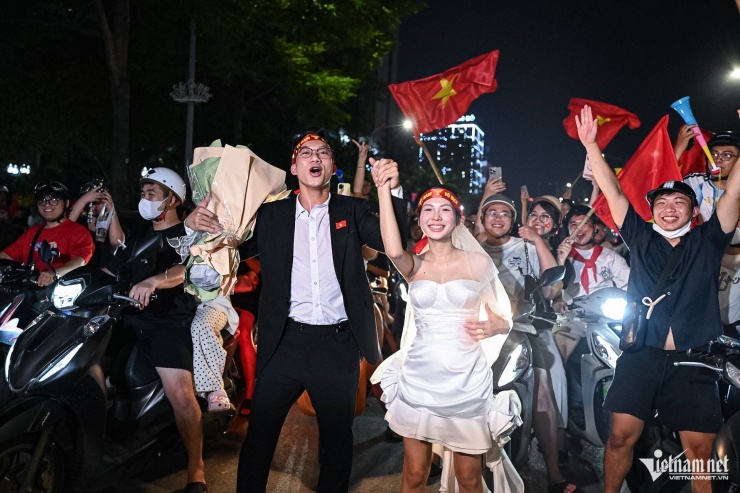 Tại khu vực hầm Kim Liên, chú rể Khổng Tiến Anh ôm theo bó hoa lớn, nắm tay vợ xuống đường ăn mừng chiến thắng. Chiếc váy cô dâu tạo điểm nhấn, khiến đám đông xung quanh reo hò không ngớt.