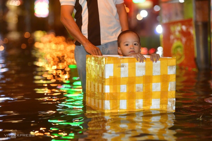 Một em nhỏ được phụ huynh đặt trong thùng xốp, đưa qua đoạn đường ngập sâu.